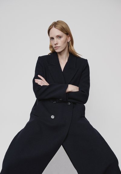 Navy double-breasted coat with large lapels, button details, and wide sleeves. Fabric shows subtle texture, offering a structured silhouette.