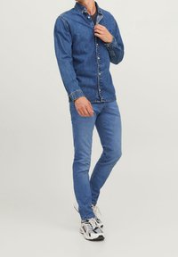 Modrá džínová košile na knoflíky s dlouhými rukávy, doplněná odpovídajícími modrými úzkými džínami a sportovními botami s barevnými akcenty.