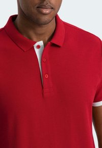 Röd polo med texturerad tyg, med en vit krage och accentränder, två röda knappar vid halsen och en figursydd design.