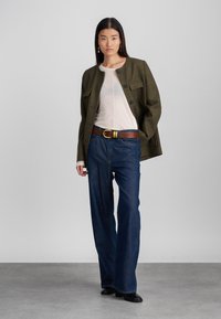 Brązowa kratkowana kurtka z przednimi kieszeniami, beżowy przezroczysty top, ciemnoniebieskie dzwony, brązowy pasek z złotą klamrą i czarne botki za kostkę.