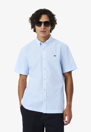 Hombre con cabello rizado que lleva gafas de sol negras, camisa de manga corta azul claro con botones y un pequeño logo verde, y pantalones oscuros.