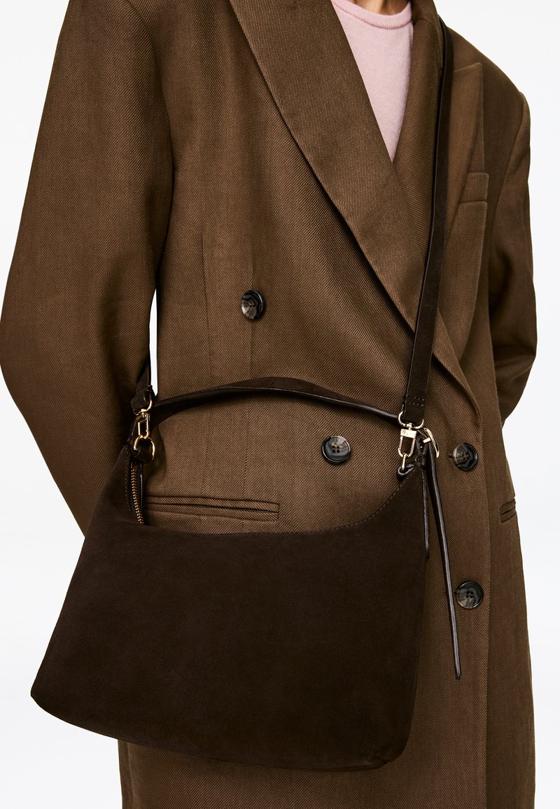 Personne portant un manteau croisé marron et un haut rose clair, portant un sac à bandoulière en daim marron foncé avec des détails dorés.