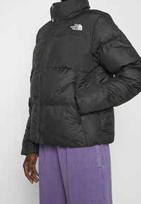 Osoba ubrana w czarną puchową kurtkę z logo The North Face i luźne fioletowe spodnie, stojąca na tle jednolitego jasnego tła.