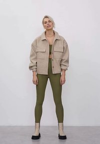 Kvinne med kort blondt hår iført beige oversized jakke, olivengrønn crop top og leggings, samt beige chunky støvler, som står mot en hvit vegg.
