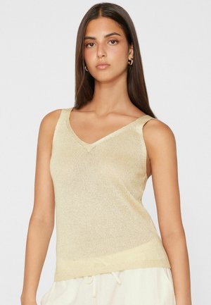 Junge Frau mit langen dunklen Haaren, die ein ärmelloses beiges Stricktop und weiße Hose mit Kordelzug vor einem schlichten hellen Hintergrund trägt.