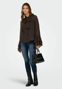 Femme portant un chemisier transparent sombre à col noué et manches à volants, jean slim bleu, bottes noires, tenant un sac à main noir à motif texturé.
