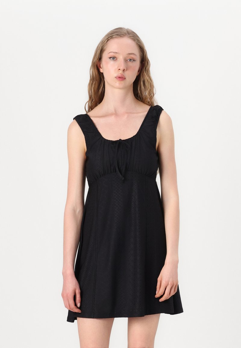 Vestido preto sem mangas com decote quadrado, alças com frufru, tecido texturizado e um laço ajustável no busto. Forma em A, com comprimento até ao joelho.