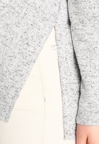 Grauer Strickpullover mit einer strukturierten Oberfläche und seitlichem Schlitzdesign. Der Stoff wirkt weich und warm, zusammen mit weißen Denim-Hosen.