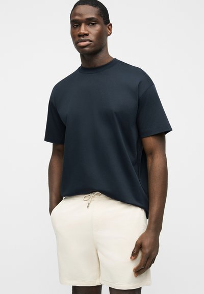 Homme portant un t-shirt oversize bleu marine uni associé à un short beige clair à cordon, debout avec une main dans la poche.