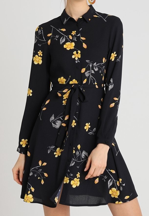 Robe noire boutonnée avec manches longues, imprimé floral jaune, col et ceinture nouée à la taille, portée par une personne debout.