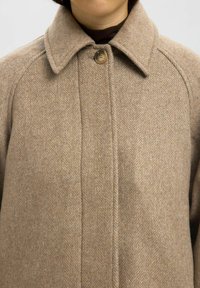 Bež herringbone plašč s pokončnim ovratnikom, zapiranjem na en gumb in teksturno volneno tkanino. Ima raglan rokave in raven kroj.