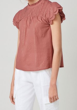 Femme portant un chemisier couleur rouille avec des manches courtes festonnées et un pantalon blanc avec poches cargo sur fond uni.