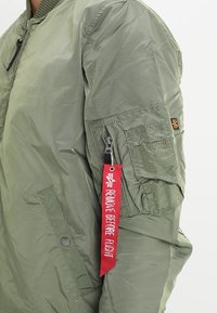 Veste bomber verte avec une poche zippée sur la manche gauche, tissu texturé et une étiquette rouge "ENLEVER AVANT LE VOL".