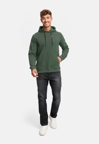 Grøn hætte sweatshirt med snørebånd, kengurulomme og ribbede manchetter, parret med mørk jeans og hvide sneakers. Modellen står.