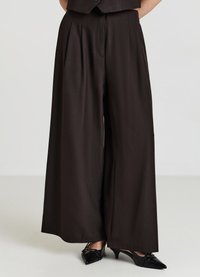 Pantalons à jambes larges en tissu marron foncé, avec des plis avant et une texture lisse. Associés à des escarpins noirs à bout pointu.