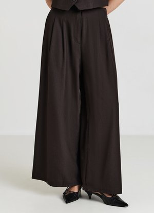 Pantaloni a gamba larga in tessuto marrone scuro, caratterizzati da pieghe frontali e una texture liscia. Abbinati a tacchi neri a punta.