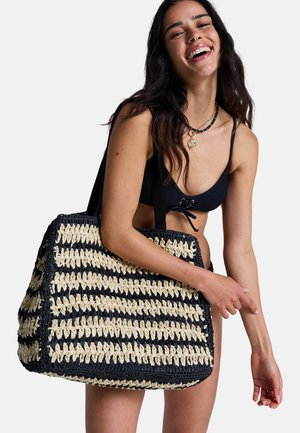 Mujer con bikini negro sosteniendo un gran bolso tote trenzado de rayas negras y beige con correas para el hombro, sonriendo contra un fondo blanco.