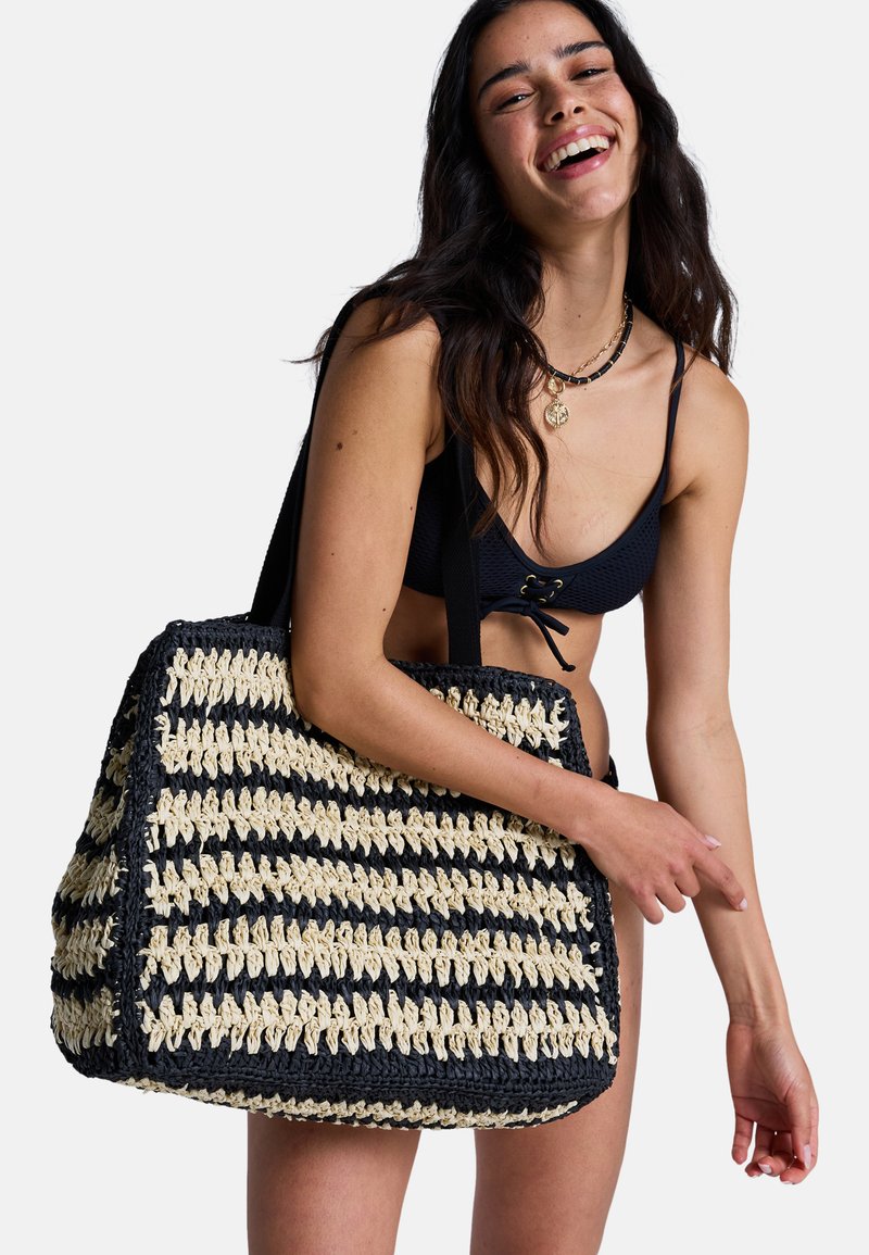 Femme en bikini noir tenant un grand sac fourre-tout rayé noir et beige tressé avec des bretelles, souriant sur fond blanc.
