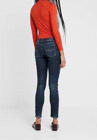 Röd långärmad topp, mörkblå slim-fit jeans med bakfickor och bruna slip-on skor. Slätt tyg med lätt stretch och en ren design.