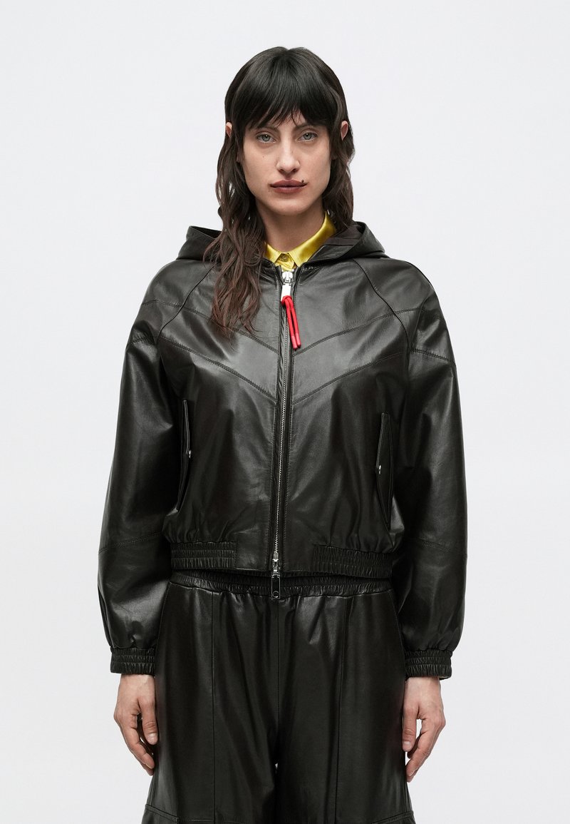 Žena s tmavými vlasmi má na sebe čiernu koženú bundu s kapucňou a nohavice, vidieť žltý golier košele, stojí pred jednoduchým svetlým pozadím.