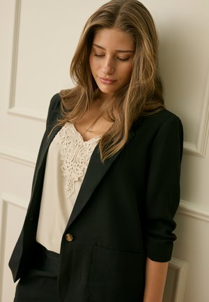 Giovane donna con capelli lunghi che indossa un blazer nero sopra una maglia crema con rifiniture in pizzo, in piedi e con lo sguardo rivolto verso il basso, davanti a una parete chiara con pannelli.