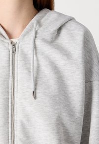 Svijetlosiva dukserica s patentnim zatvaračem izrađena od meke, teksturirane tkanine. Ima kapuljaču, srebrni patentni zatvarač i vezice s metalnim vrhom.