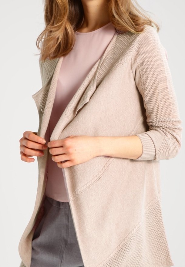 Femme portant une chemise rose clair, un cardigan beige texturé ouvert devant et un pantalon gris, ajustant le cardigan avec les mains au niveau de la taille.
