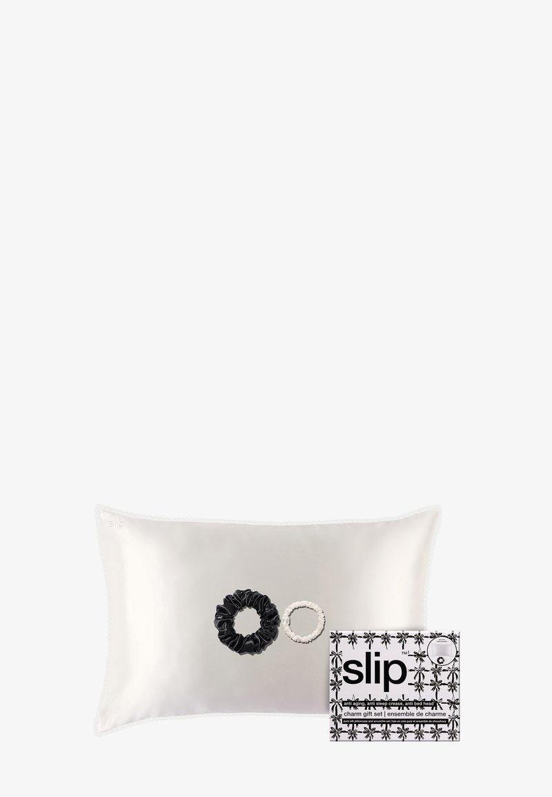 Silkkatyyny vaaleassa kermassa, jonka edessä on musta-valkoiset hiuspidikkeet. Mukana kukkakoristeilla varustettu lahjapakkaus.