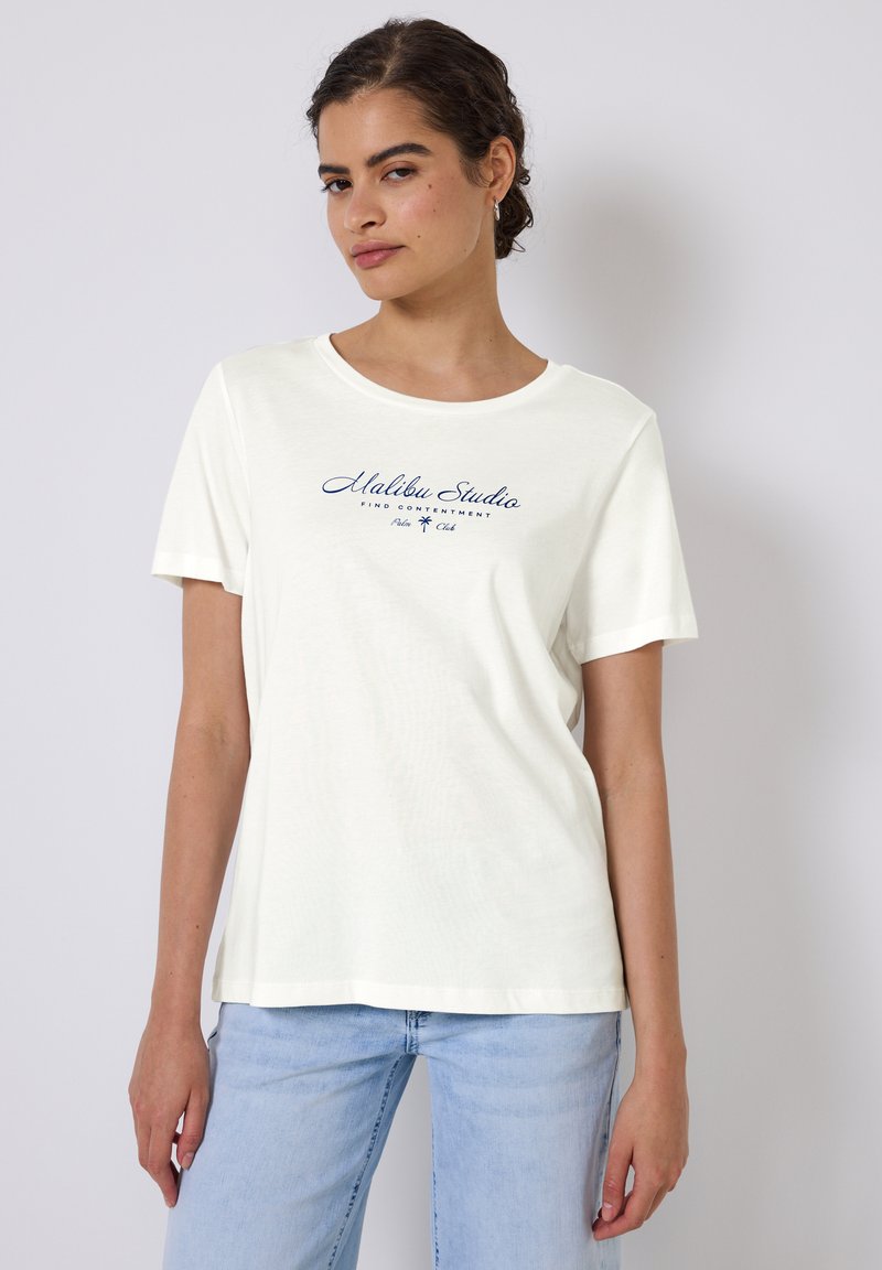 T-shirt crème à manches courtes en tissu doux, ornée d'une impression bleu marine avec le texte « Malibu Studio » et une icône de palmier.