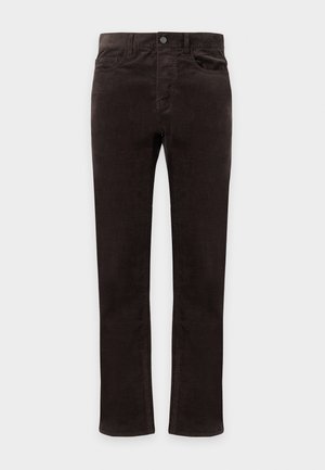 Homme debout portant une chemise à manches longues foncée, un pantalon noir, des bottes noires et une ceinture avec une boucle argentée sur un fond blanc uni.