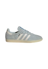 Světlé šedé tenisky Adidas Samba s bílými pruhy, s gumovou podrážkou a textem "SAMBA" na boční straně, viděné z vnější strany.
