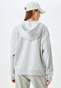 Felpa grigia con cappuccio, dalla vestibilità relax, con una consistenza morbida, polsini e orlo a costine, abbinata a pantaloni jogger grigi chiari e un cappello beige.