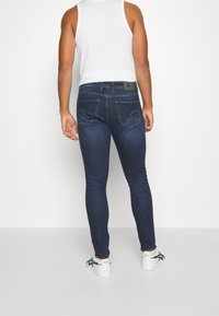 Smal passform mörkblå jeans med en lätt tvätt, fem fickor och en svart etikett vid midjan. Bärs med en vit linne och sneakers.