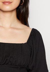 Svart top med fyrkantig halsringning och samlad tygdetalj, med korta puffärmar och en texturerad yta, mot en neutral bakgrund.