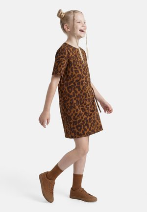 Smiling girl with blonde hair in two braids and a bun, wearing a brown leopard print dress, brown socks, and brown sneakers, walking right.