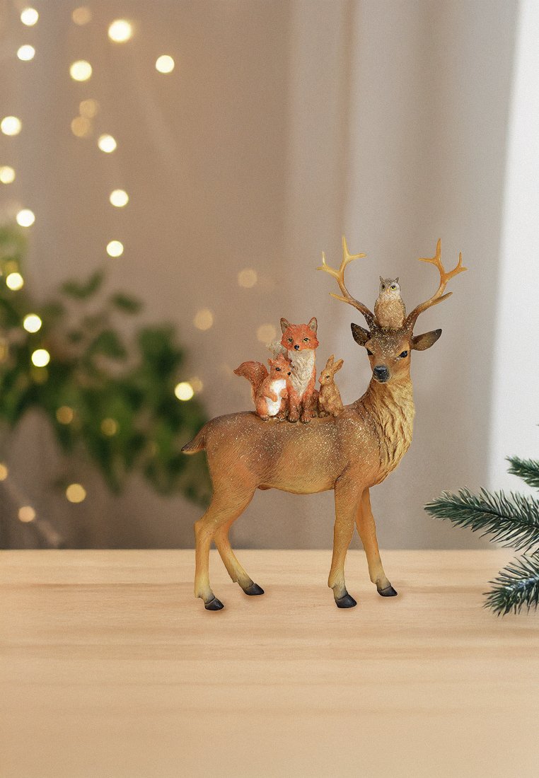 Figura di cervo con corna che tiene un gufo, una volpe, uno scoiattolo e un coniglio sulla schiena, posizionata su un tavolo di legno con uno sfondo di luci sfocate.