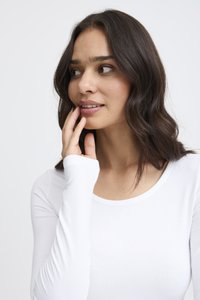 Valkoinen pitkähihainen paita, jossa pyöreä pääntie, valmistettu pehmeästä kankaasta. Mallilla on aaltoilevat tummat hiukset, ja hän koskettaa hellästi kasvojaan.