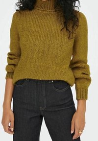 Olivengrüner Strickpullover mit hohem Kragen, geripptem Textur und leicht lockeren Ärmeln; kombiniert mit dunkelblauen Jeans.