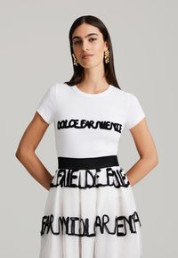 T-shirt en coton blanc avec des lettres texturées noires. La jupe présente des couches de tissu blanc transparent avec des accents de texte noir frangé et une taille noire.