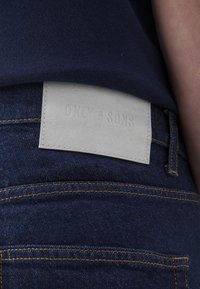 Mørkeblå denimjeans med et lysegrått lærmerke med preget tekst, plassert på livet, som viser oransje sømdetaljer.