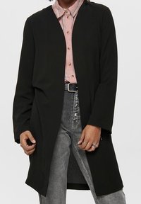 Długi czarny kardigan o luźnym kroju, wykonany z miękkiego materiału. Posiada otwarty przód, długie rękawy oraz gładką strukturę. Zestawiony z koszulą na guziki.