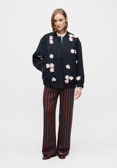 Jachetă bomber neagră cu aplicații florale roz; are guler și manșete ribbed. Asortată cu pantaloni cu dungi verticale roșii și negre.