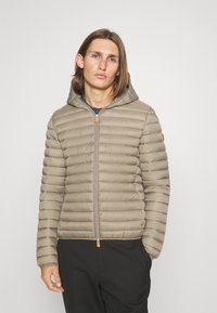 Beige puffer jacket with horizontal quilted pattern, attached hood, and orange zipper pull. Pockets at the sides, smooth fabric texture.