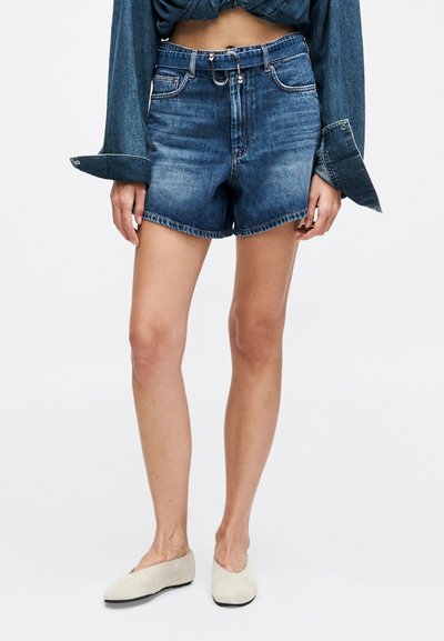 Μοντέλο που φοράει μπλε τζιν σορτς μεσαίου ύψους με γυρισμένα τελειώματα, συνδυασμένα με δεμένο μπλε τζιν πουκάμισο και μπεζ παπούτσια slip-on, στέκεται μπροστά σε λευκό φόντο.