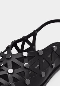 Μαύρο σανδάλι με γεωμετρικό σχέδιο με περικοπές και στρογγυλές μεταλλικές καρφίτσες στο πάνω λουρί, ανοιχτή φτέρνα και επίπεδη σόλα.