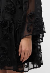 Crna prozirna haljina od tkanine s teksturiranom cvjetnom izvezenom. Ima lepršavi dizajn, višeslojni rub i složene detalje duž rukava.
