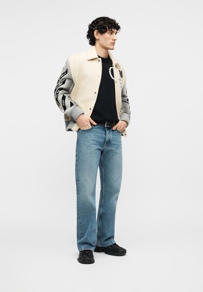 Νεαρός άνδρας με μπεζ και γκρι varsity μπουφάν, μαύρο πουλόβερ με σχέδιο καρδιάς, μπλε τζιν και μαύρα αθλητικά παπούτσια στέκεται σε ουδέτερη στάση.