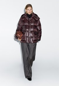 Burgundy puffer jacket with a high collar, front pockets, and glossy texture, paired with gray high-waisted pants and a brown handbag.
