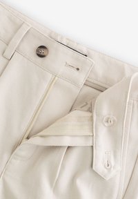 Beige nadrág gombos és cipzáras záródással, sima textúrával, övbujtatókkal és egy kis logócímkével belül.