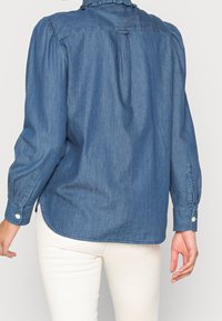 Jeanskjorta med långa puffärmen, volangkrage, detalj med ryggrynna och knäppta manschetter. Färg: mellanblå. Bärs med ljusa byxor.